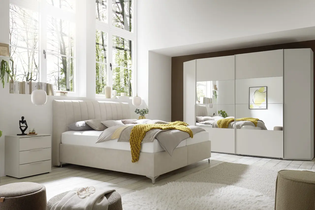 Polsterbett mit Kissen und Decke vor Fenster, Kleiderschrank mit Spiegeltüren rechts und Nachttisch links im Schlafzimmer